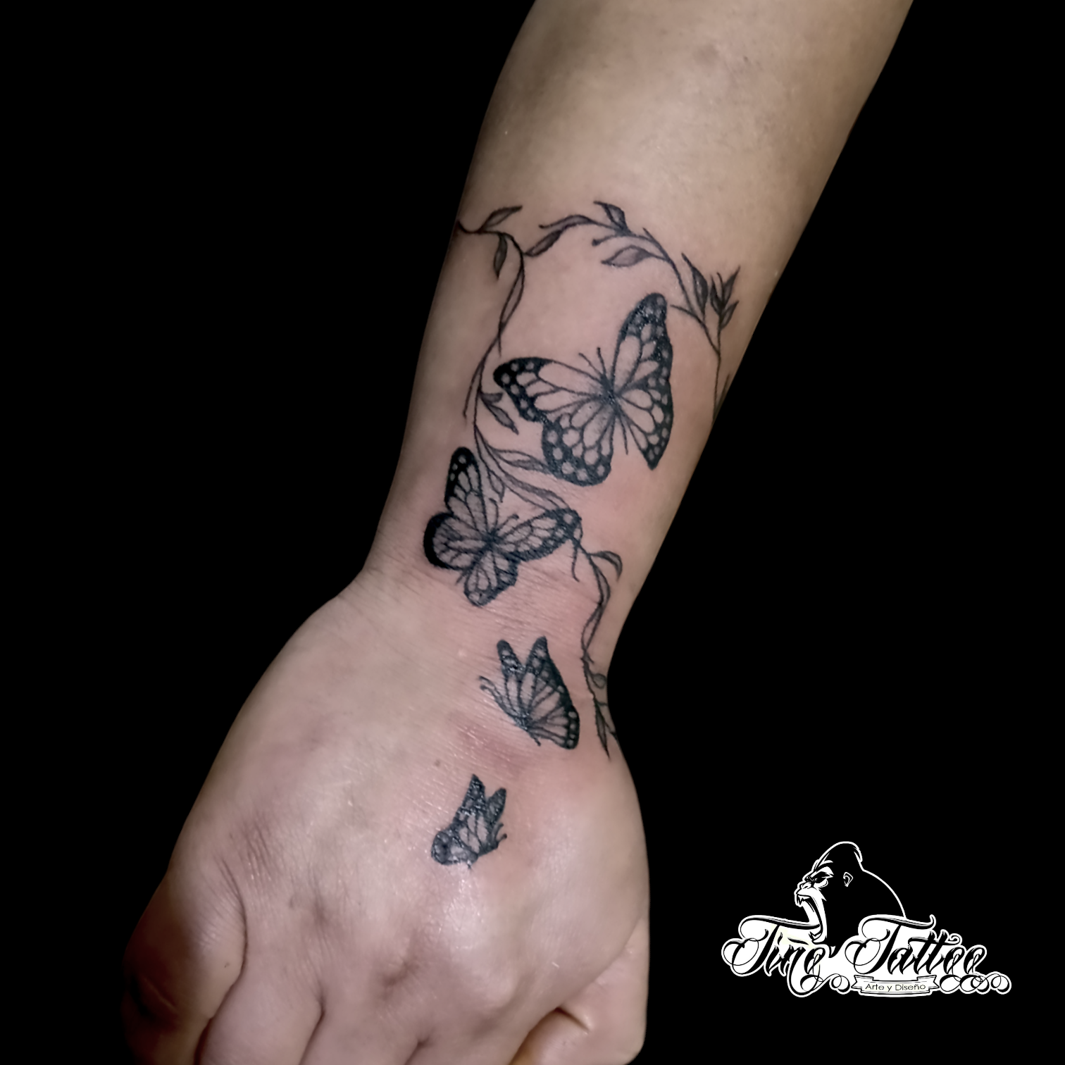 Tatuaje en negro de varias mariposas con ramas delicadas sobre la mano y muñeca