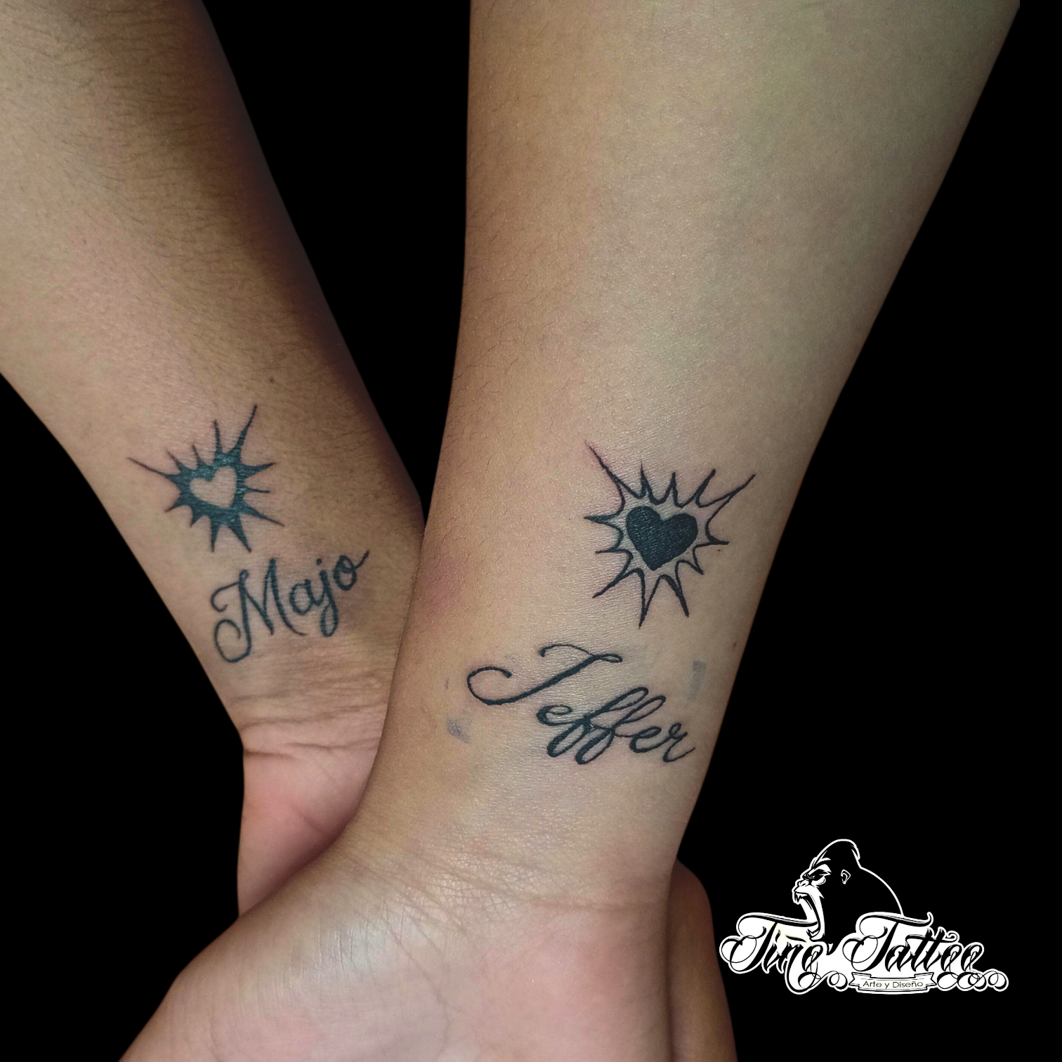 Tatuajes a juego con corazones negros y lettering con nombres en dos muñecas