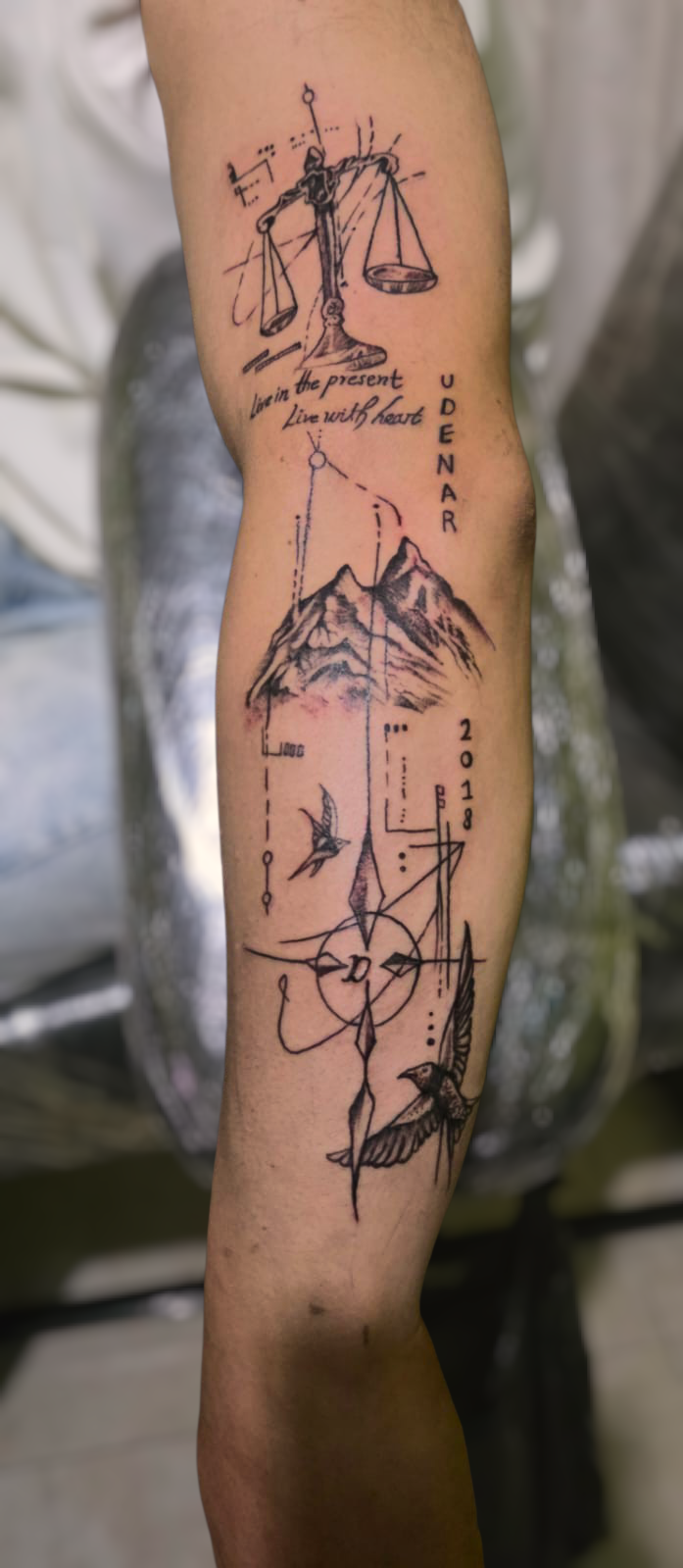 Tatuaje fineline con balanza, montañas, aves y trazos geométricos en antebrazo